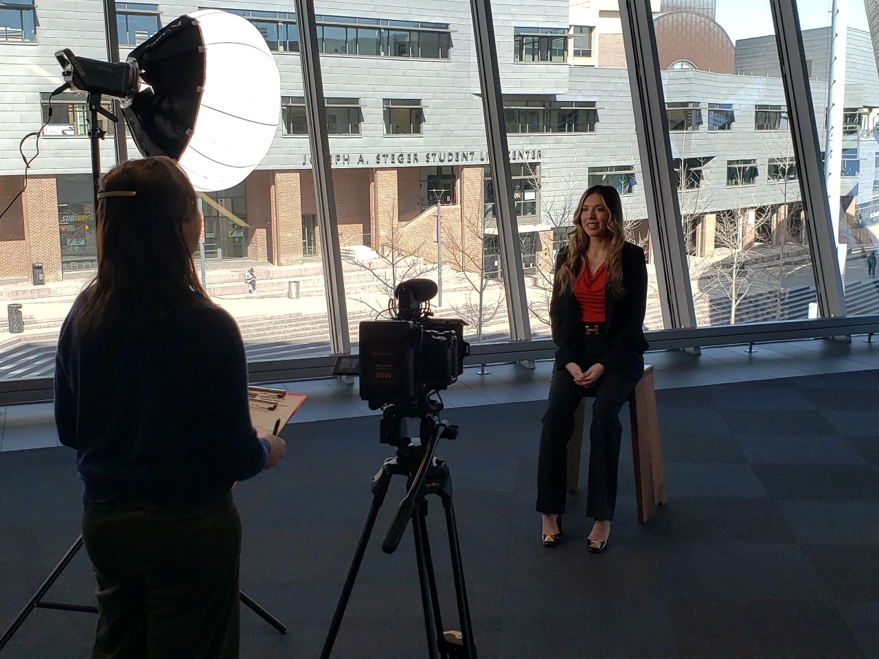 Ashley Merianos shown seated on a bench with Stegar Student Center showing in the background and a camera and light pointed at her.  We see the back of a producer who asks questions.