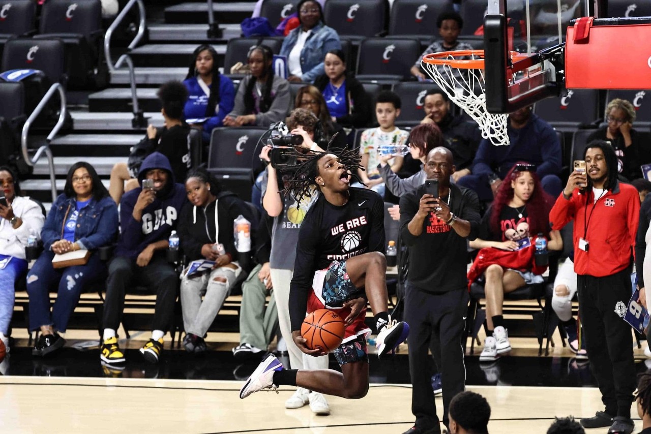 a crowd watches in Fifth Third Arena as a camera person snaps a photo of a young male basketball player running down the court to dunk the basketball