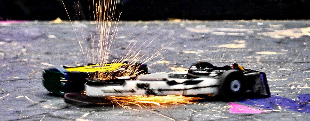 A 12-pound combat robot called Maximizer shoots sparks from its front spinning wheel while bumping against another small robot.