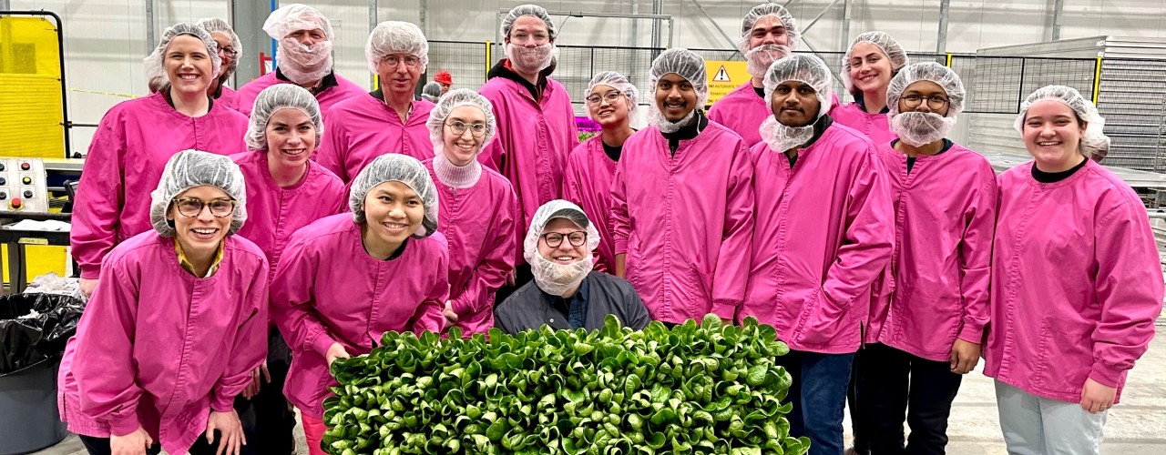 Several UC student wearing pink jackets and hair nets surround an 80 Acres employee holding a box of hydroponically grown spinach.