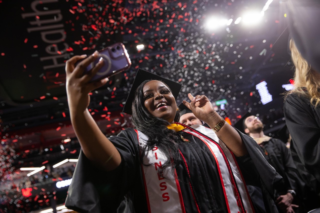 A UC grad in her cap and gown takes a selfie while holding up a peace sign as confetti rains down from the rafters of Fifth Third Arena.