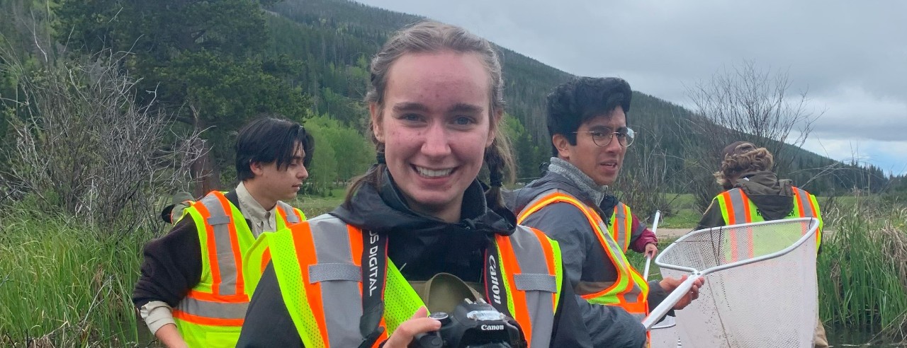 young woman outdoors holding a camera while people behind her hold large nets