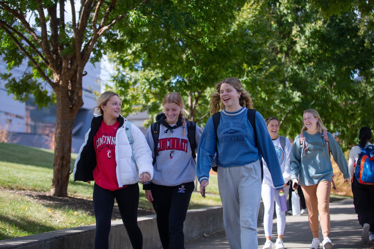 five female students all smiling and in colorful sweatshirts walk across the UC campus 