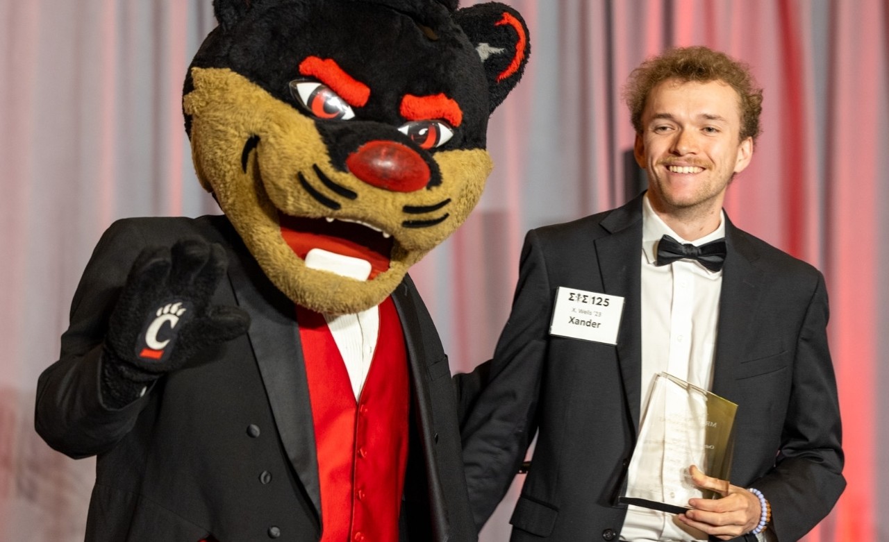 A male student in a black suit holds an award standing next to the UC Bearcat mascot.