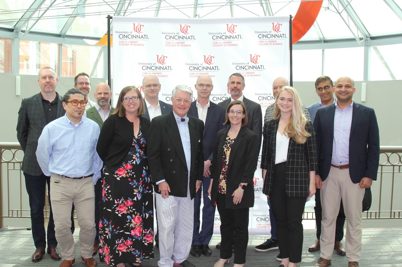 Fourteen people stand in front of a UC-branded step and repeat in an atrium