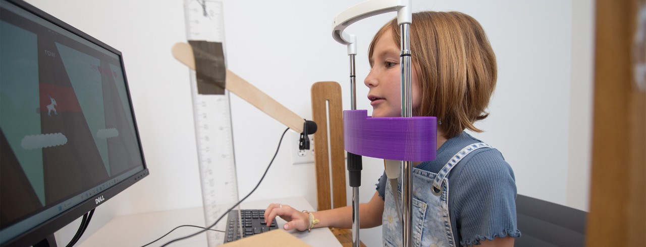 A girl with her chin on a chin rest looks at a computer screen with an animated goat on it.