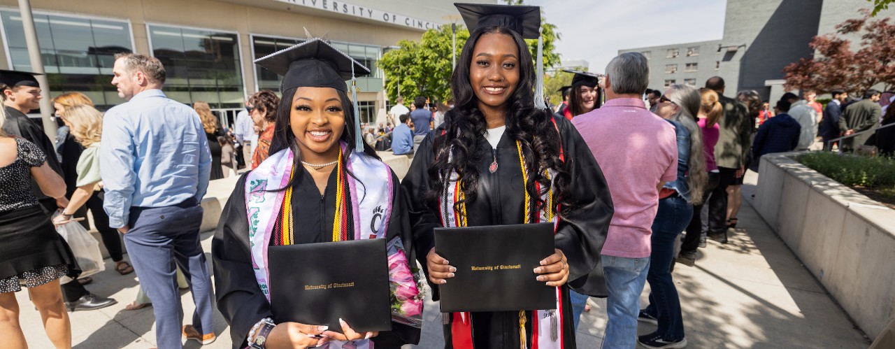 UC grads Precious Randall on left and Corin Manning on right stand outside Fifth Third Arena wearing commencement cap and gown holding their diplomas.