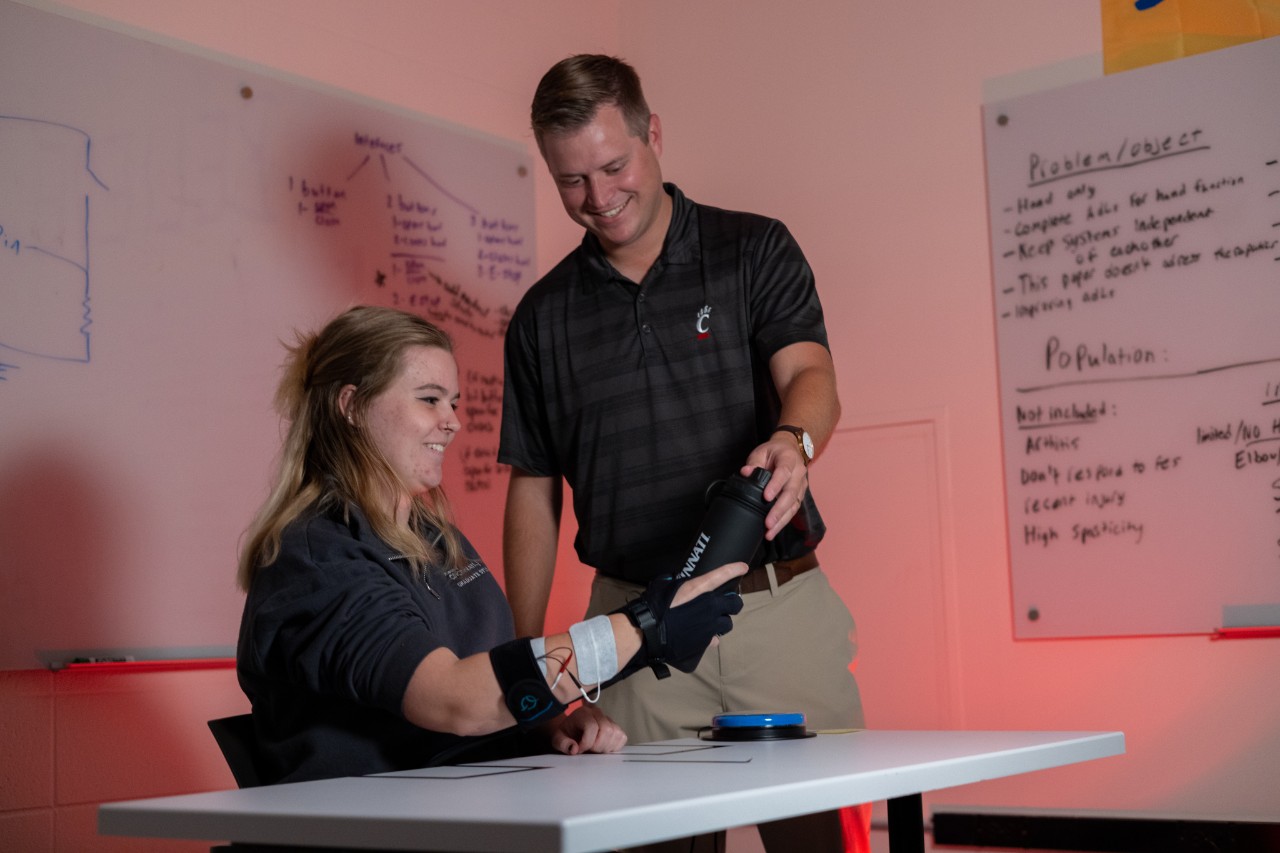 A student sits at a table wearing an exoskeleton prototype on her hand while Derek Wolf stands and examines it
