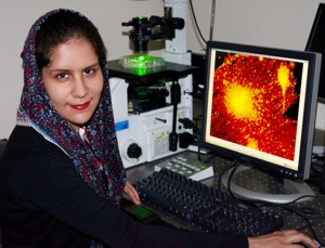 Woman sits next to a cumputer screen with a magnified organic cell illuminated on the screen.