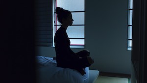A teenage girl sits cross-legged on a bed in a darkened room.