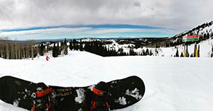 A view of a snowy, tree-lined ski slope on a bright day, taken from a point of view that shows the photographer's feet in a snowboard. The skies are blue with clouds and there is only one other person on the slope.